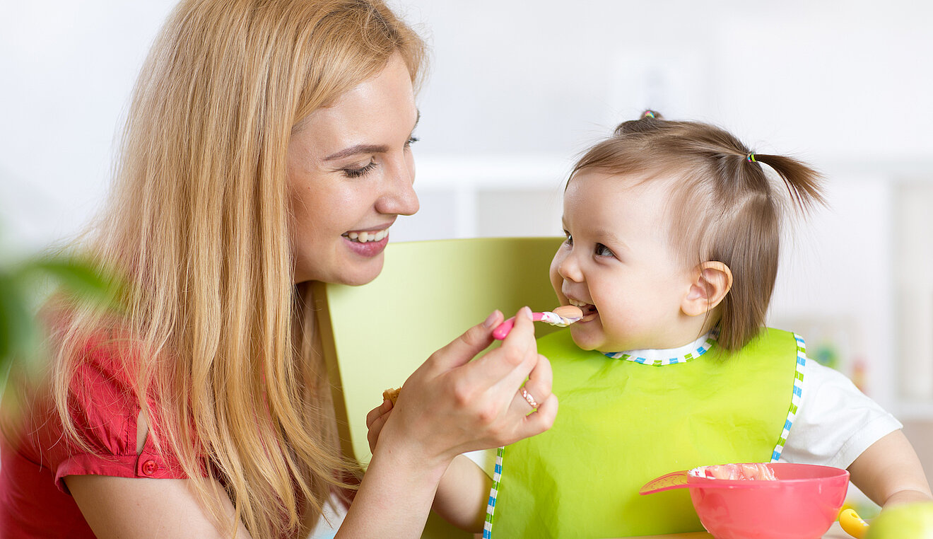 Baby sitzt im Hochstuhl und wird von der Mutter mit einem Löffel gefüttert.