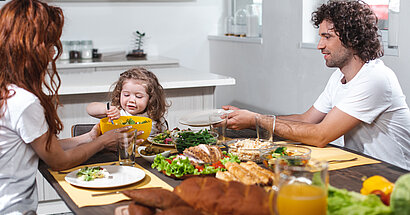 Familie sitzt am Esstisch und isst. Vater lässt sich vom Kind den Teller füllen.