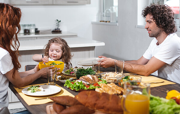 Familie sitzt am Esstisch und isst. Vater lässt sich vom Kind den Teller füllen.