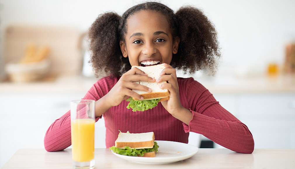 Junges Mädchen sieht lächelnd in die Kamera und beißt in ein Sandwich