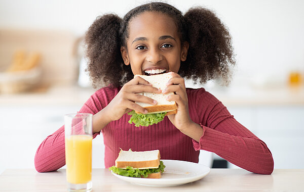 Junges Mädchen sieht lächelnd in die Kamera und beißt in ein Sandwich