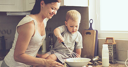 Junge, KitaKind, rührt Kuchenteig in einer Schüssel. Sohn sitzt auf Arbeitsfläche und Mutter steht vor ihm und passt auf.