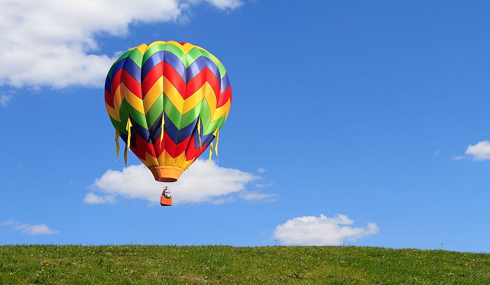 Bunter Heißluftballon schwebt auf eine grüne Wiese zu.